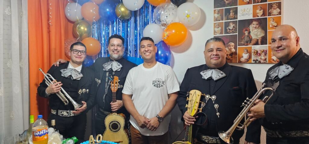 Mariachis con instrumentos junto a un invitado ante una decoración de globos azules y amarillos.