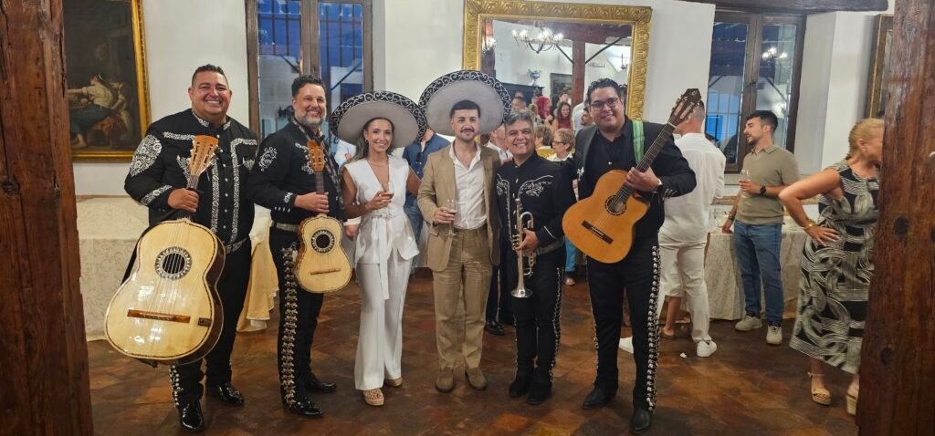 Mariachis con guitarras y trompetas posan junto a dos invitadas durante una celebración en un salón.