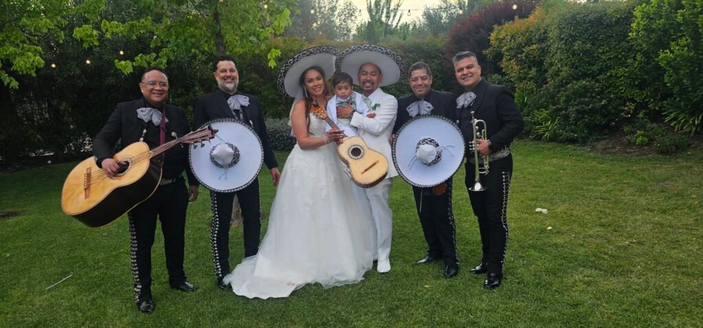 Grupo de mariachis con sus instrumentos junto a una pareja de novios en un jardín.