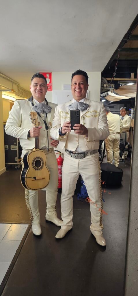 Dos músicos con trajes de charro claros sostienen una guitarra y una trompeta.